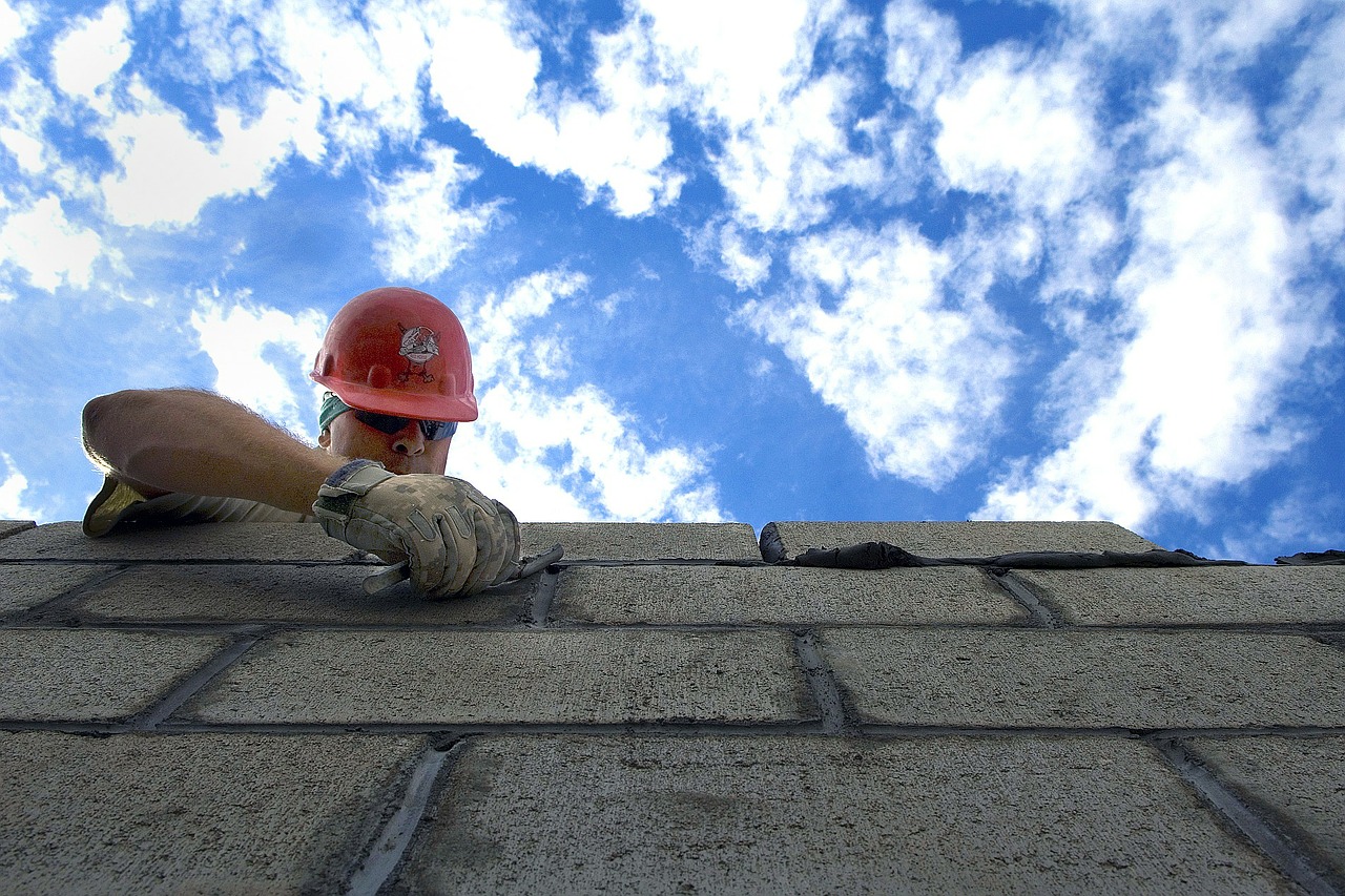 photo of a man in an orange helmet building a brick wall against a partly cloudy blue sky