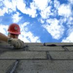 photo of a man in an orange helmet building a brick wall against a partly cloudy blue sky
