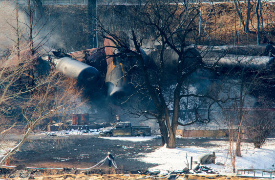 derailed oil cars behind a vacant lot with a pickup truck and charred trees