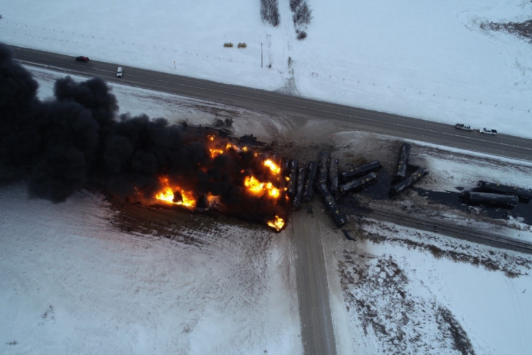 charred remnants of oil cars on fire in the snow next to roadway