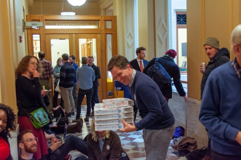 Group of people waitin in line in Portland City Hall. At the center of the image, a 40-year-old man in a pullover sweater shares a laugh while distributing pizza.