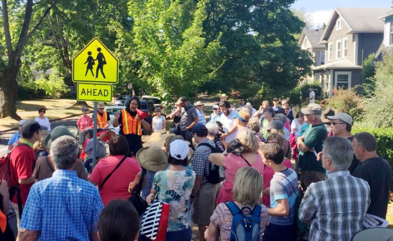 A group of more than 30 people listen to a woman on a city street in a tree-lined neighborhood