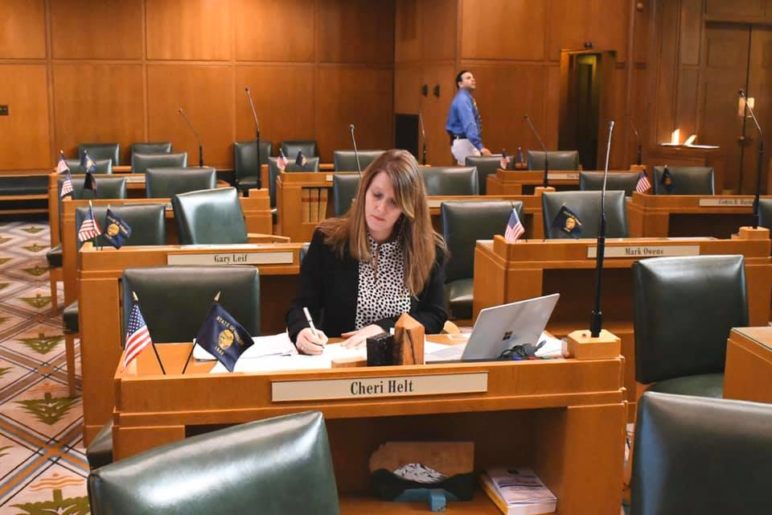photo of Rep. Cheri Helt, a woman in early middle age, working at a desk in the state legislative chambers. All other desks around her are empty.