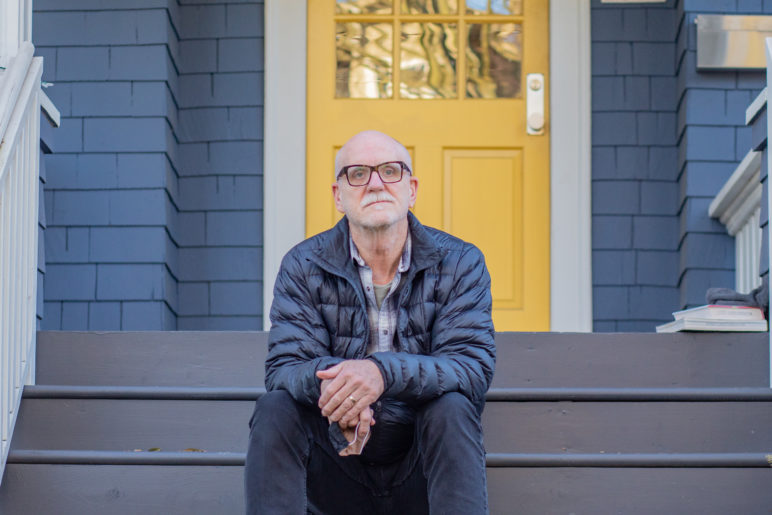 Profile photo of Joe Zehnder sitting on the steps of his blue house with a yellow porch