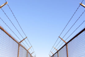 A view of barbed chain linked fences on either side of the viewer, under a blue sky.