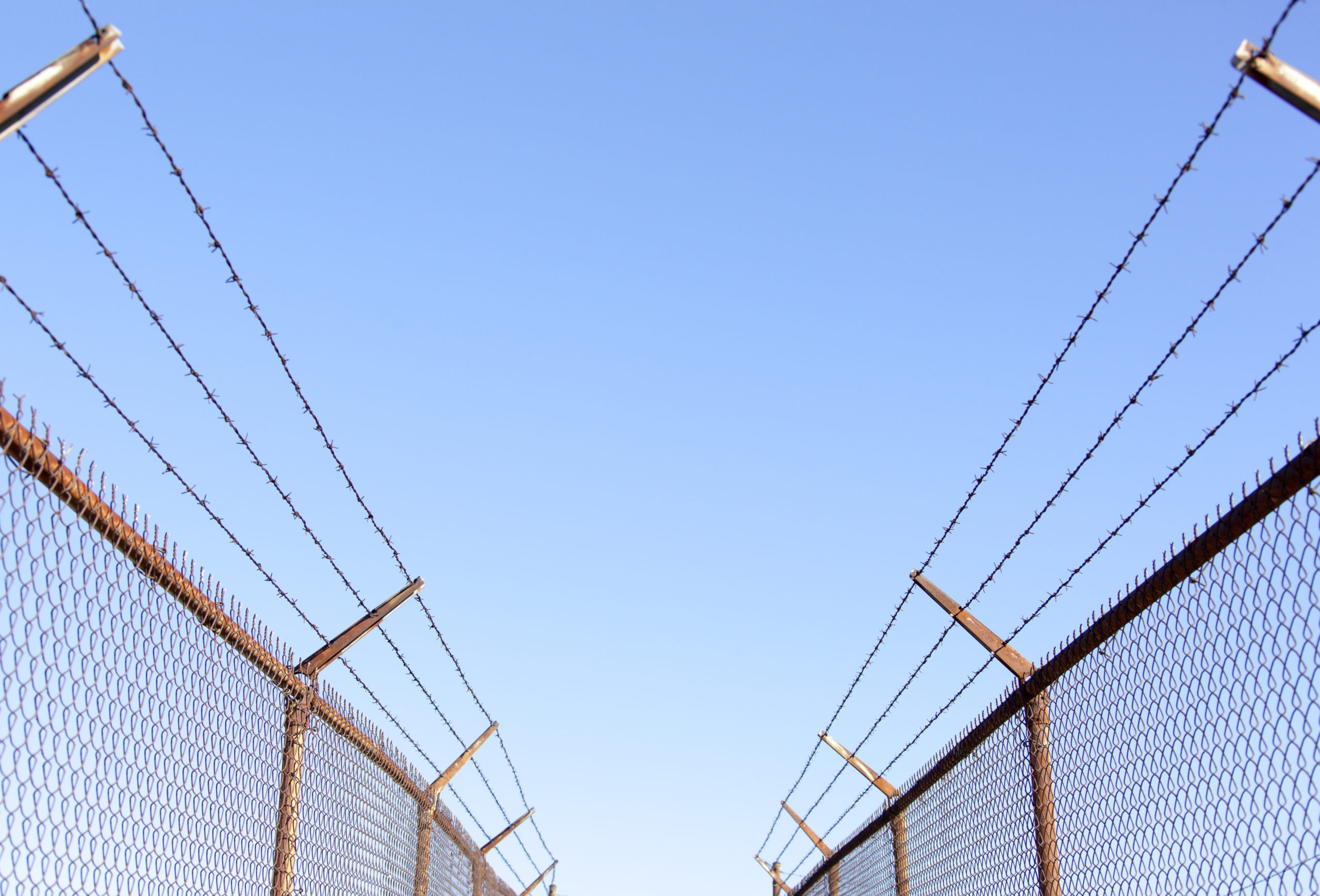 A view of barbed chain linked fences on either side of the viewer, under a blue sky.