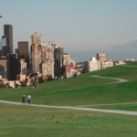 Seattle skyline, looking south toward the buildings of downtown, with Mount Rainier on the horizon and people walking the trails of the Seattle Art Museum sculpture park in the foreground.