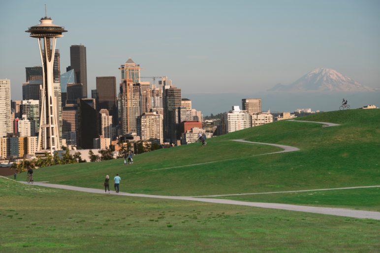 Seattle skyline, looking south toward the buildings of downtown, with Mount Rainier on the horizon and people walking the trails of the Seattle Art Museum sculpture park in the foreground.