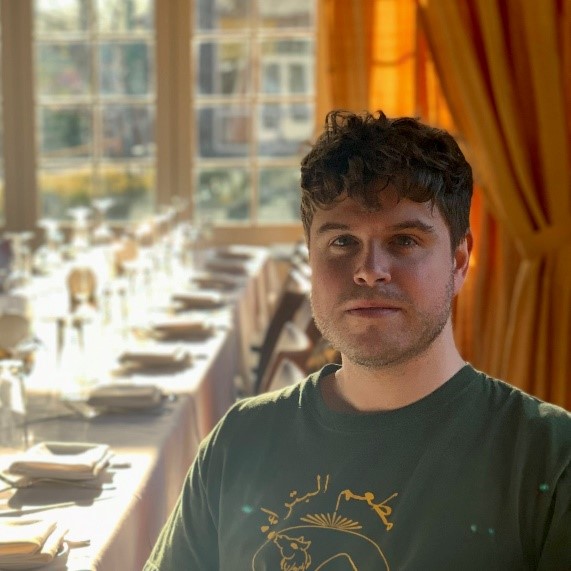Photo of Brandon Rostek of the Atlas the Restaurant. Young man in a green shirt standing before a banquet room.