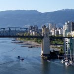 Photo of the Burrard Bridge in Vancouver, BC with the right side of the photo showing many tall skyscrapers along the waters and a mountain backdrop