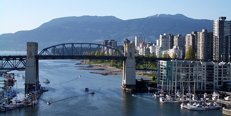 Photo of the Burrard Bridge in Vancouver, BC with the right side of the photo showing many tall skyscrapers along the waters and a mountain backdrop
