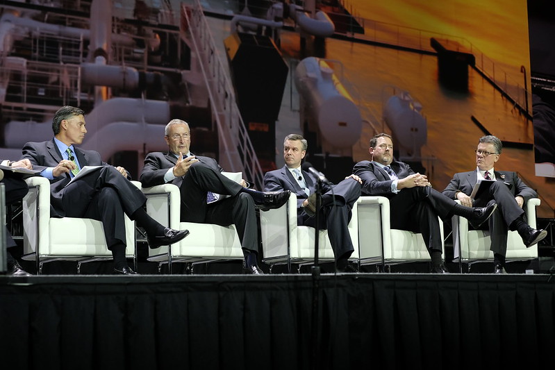 Five white men sitting in business suits in white lounge chairs on stage in front of a screen depicting gas pipes