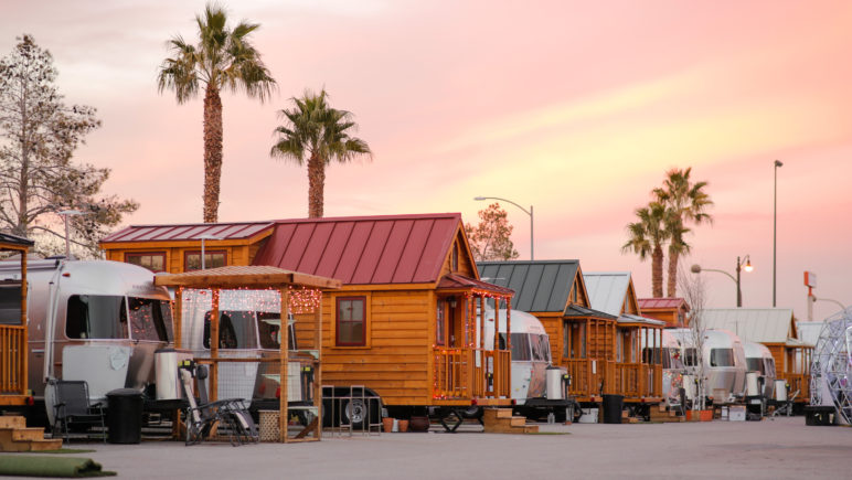 A row of airstream trailers and wood-paneled tiny houses on wheels in a smooth, undeveloped lot. In the background, a pink sunset sets of several palm trees.