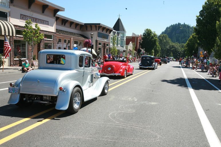 A row of vintage cars rolls down a two-story downtown main street as people look on from the curbside areas. Buildings on the main street do not have any off-street parking.