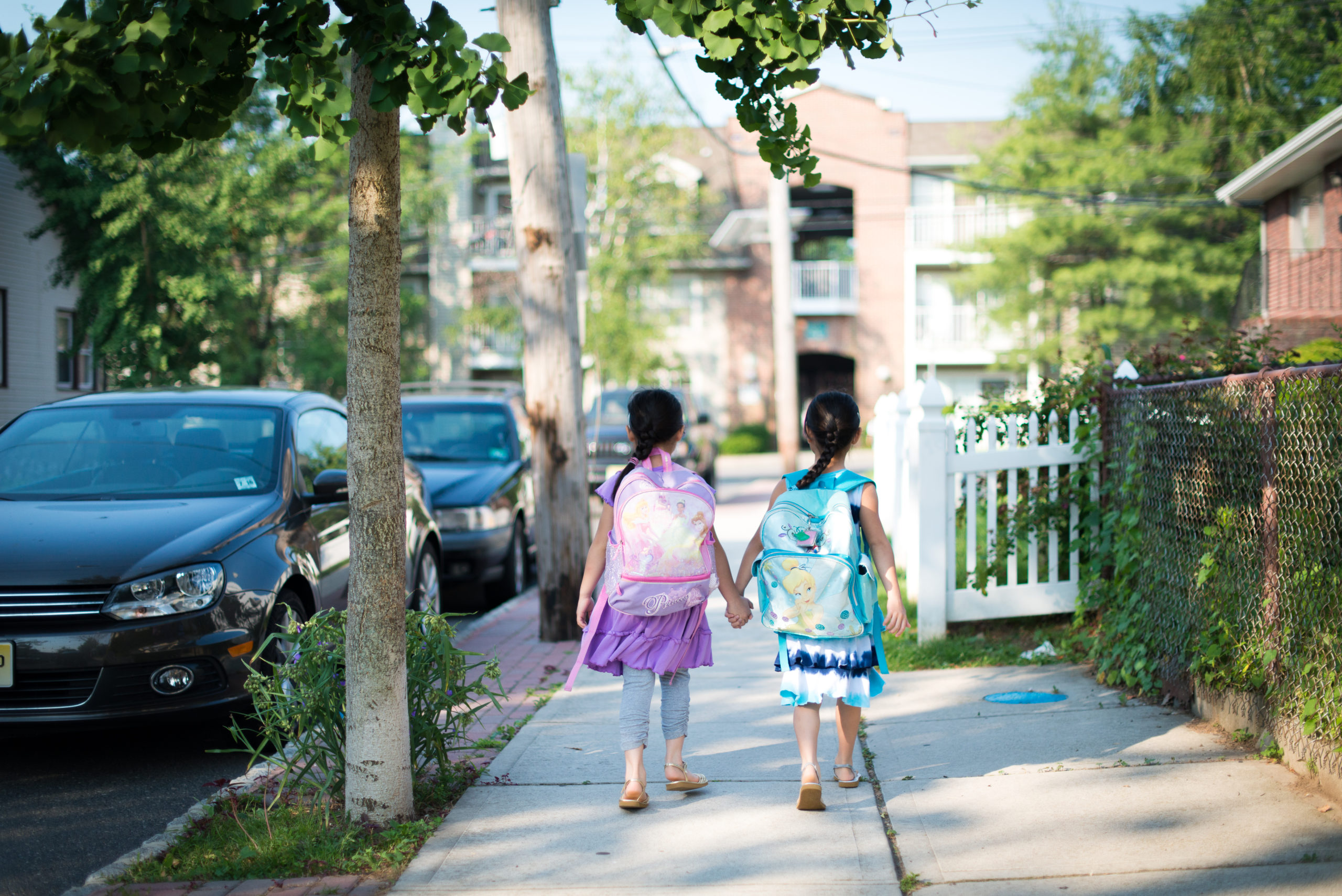 Two young children walk on a sidewalk in a city neighborhood, wearing school backpacks and holsing hands. There are trees and lush greenery along the road and sidewalk. An apartment building is directly ahead of them. There are cars parked on the street to their left. It's a sunny day.