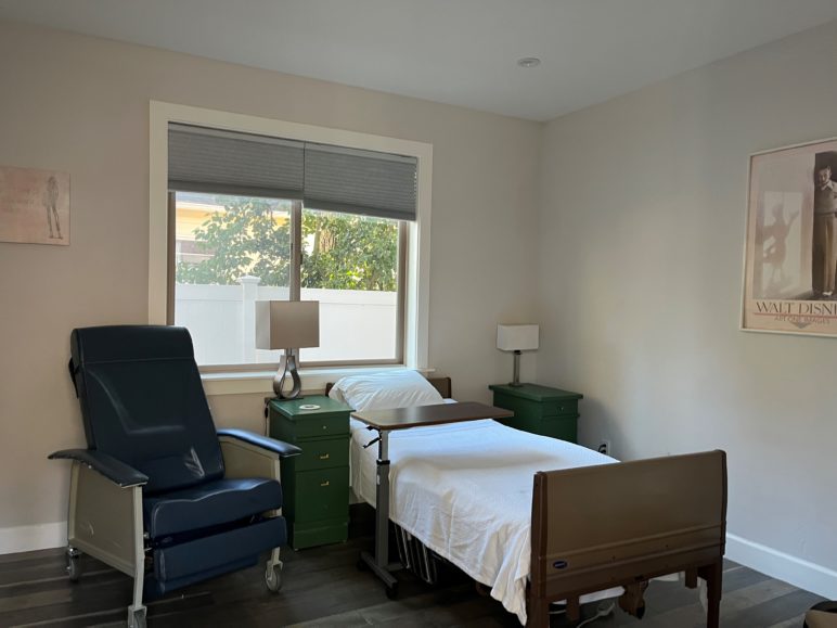 An empty room at Rooted Care Communities. Single bed with white linens next to nightstands and a black armchair. Photo by Catie Gould. 