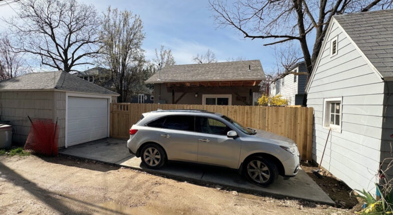 a photo of a car on a parking pad between a home and a garage, alongside a high fence