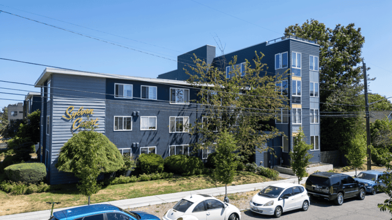 Photograph by Alex Hart Photography. Apartment buildings alongside a sunny city street. Lots of trees and green topiary, with windows alongside each unit.