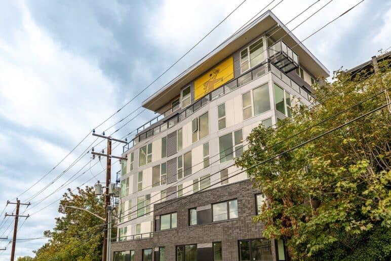 Photograph courtesy of BUILD LLC. Photo of a multi-unit building with windows covering nearly the entirety of the sides. A deck area can be seen at the top, just above powerlines long the street.