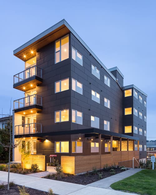 Photograph courtesy of Schemata Workshop. A multi-story apartment building at dusk, with lights on. Large pane windows and decks can be seen on the side.