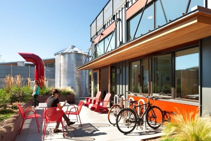 Resident common area. Photograph courtesy of Lara Swimmer Photography. Large pane windows on a building, red lawn furniture and bike parking (with bikes) can be seen on a sunny day. A refurbished silo can be seen in the background.