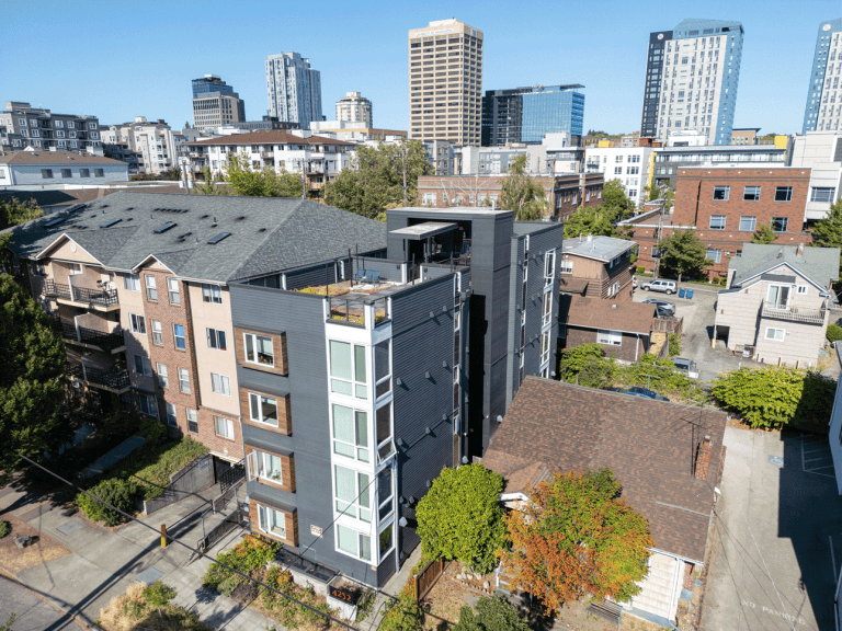 Photograph by Alex Hart Photography. A 4 or 5-story multi-unit building can be seen in a narrow city lot, next to single-unit houses on equal (or larger) sized lots. City scape can be seen in the background on a sunny day.