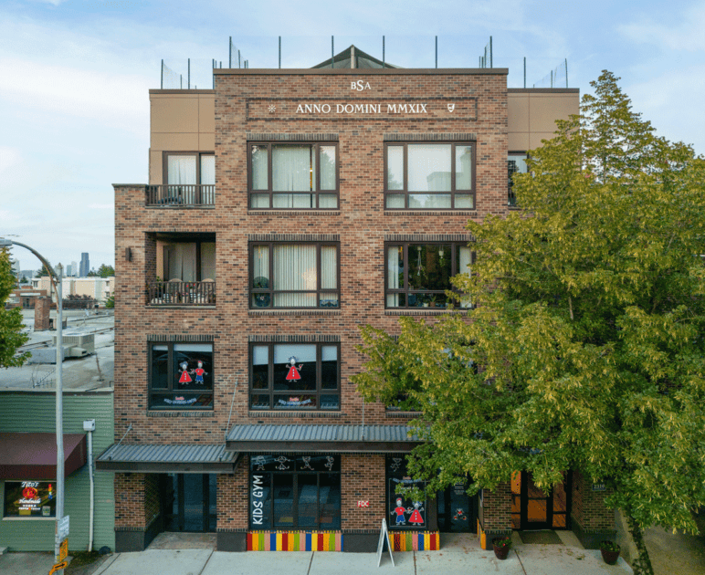 Photograph by Alex Hart Photography. A four-story brick building with large windows to each unit. Decks railings can be seen, and a children's store/gym can be seen on the ground floor. A large tree frames the side.