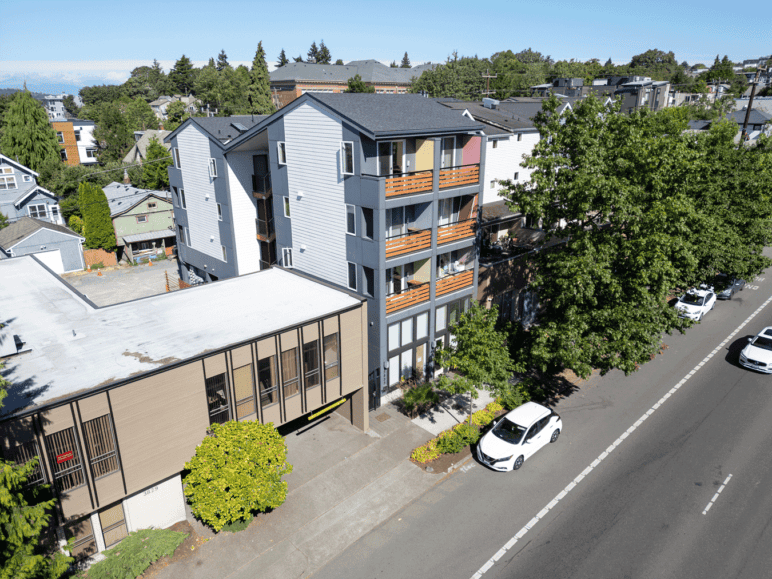 Photograph by Alex Hart Photography. Aerial view of a 4-story apartment building on the street, fitting alongside other smaller buildings on same-sized lots.