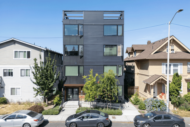 Photograph by Alex Hart Photography. Simple 4-story muti-unit building sits in between single-unit houses on a same-sized city lot. Large windows and a open rooftop deck can be seen.
