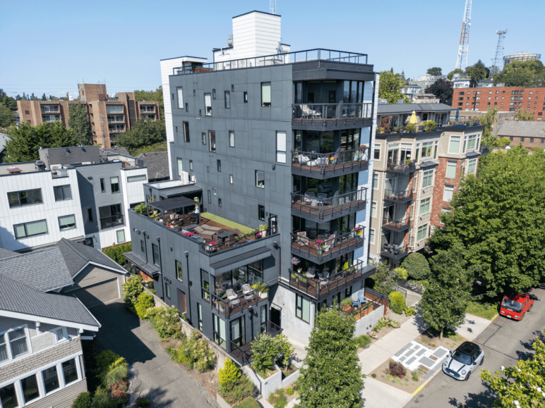Photograph by Alex Hart Photography. Aerial of some apartment buildings, focusing on a 6-story building with decks on a sunny day. A joined common-area building can be seen with what looks like a rooftop garden.