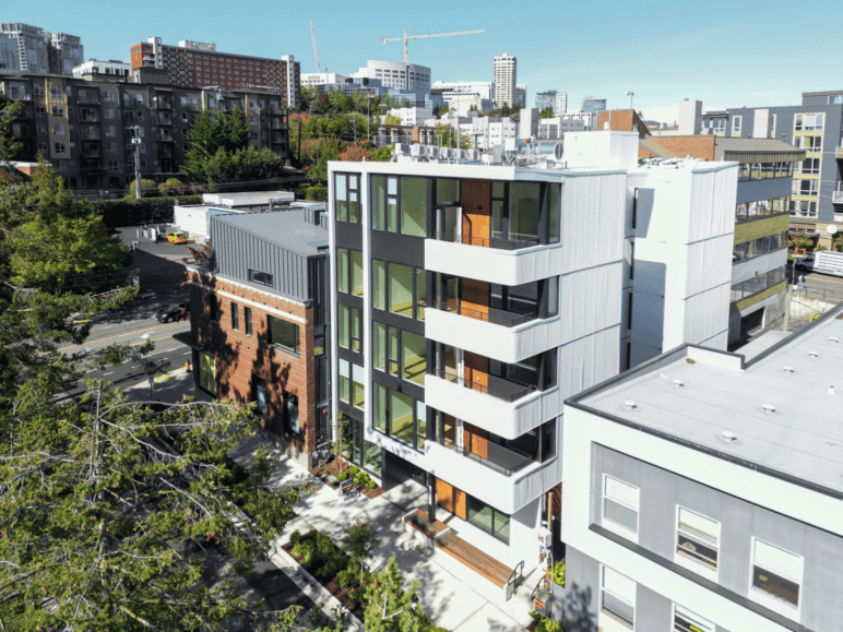 Photograph by Alex Hart Photography. Tall 5-story building can be seen. Large windows along each floor and the stairwell, as well as private decks to each unit on a sunny day in the city.