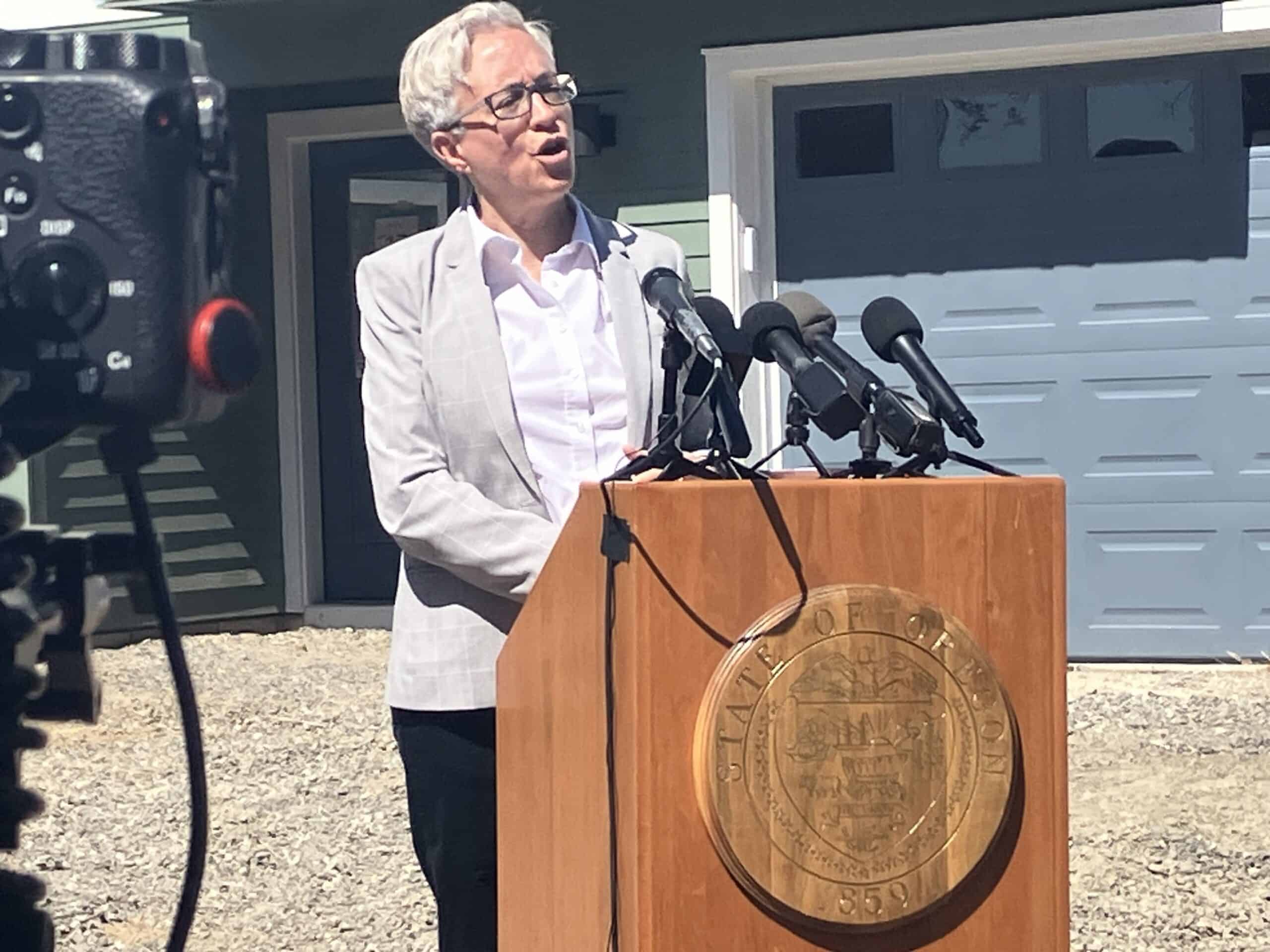 A white woman with white hair and a light gray suit jacket stands behind a lectern with the seal of the State of Oregon on it. It is set up on dirt and gravel outside a newly built home.