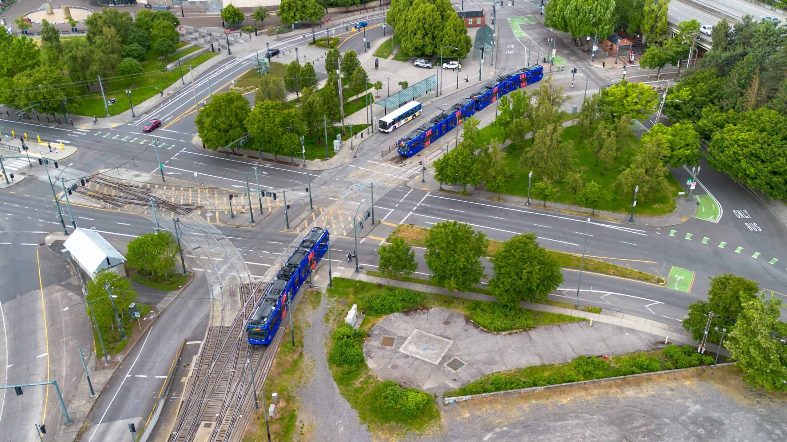 Portland, Oregon, USA - May 18, 2025 – Aerial view of two blue Trimet MAX Trains and a Trimet bus, plus security vehicles and private autos, winding through small, tree-filled city blocks.
